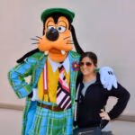 Woman posing and smiling with a person in a Goofy costume wearing a colorful plaid suit, outdoors against a plain background.