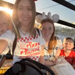 A woman and three smiling children sit together in a brightly lit gondola ride, sunlight streaming in through the window behind them.