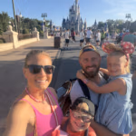A smiling family of four poses for a selfie at a theme park, with Cinderella’s Castle visible in the background.
