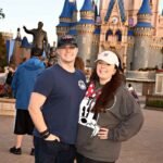 A smiling couple poses in front of Cinderella's Castle at a Disney park, wearing casual clothes and hats, with other visitors and a statue in the background.