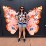Woman standing in front of colorful butterfly wings mural at Epcot International Festival of the Arts, smiling and holding a drink.