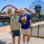 Two men posing and smiling at Disney California Adventure’s Pixar Pier, with the Incredicoaster and Mickey’s Fun Wheel in the background on a sunny day.